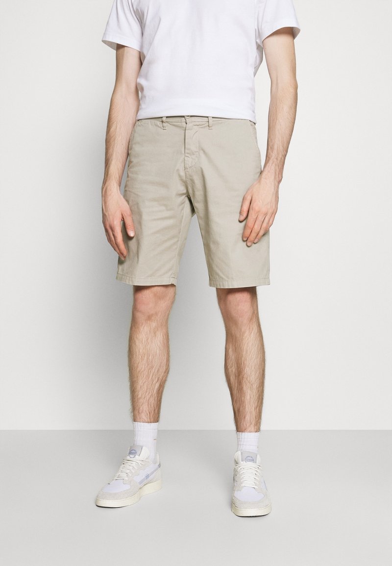 Shorts beige clair en coton, avec une taille plate et des poches latérales. Assortis à un t-shirt blanc uni et des baskets blanches.