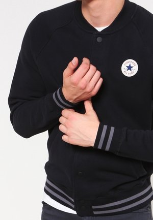 Mand iført en sort Converse-jakke med stribede manchetter, der griber sit håndled med den anden hånd mod en hvid baggrund.