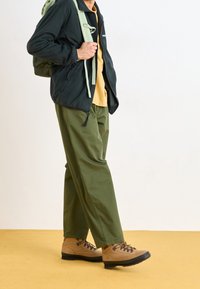 Mörkgrön zip-up jacka, beige t-shirt, olivgröna byxor och ljusbruna vandringskängor med svarta sulor. Bär en ljusgrön ryggsäck.