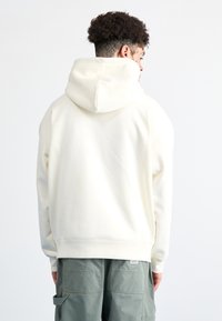 Sudadera con capucha de color beige claro, hecha de tela suave, que presenta un bolsillo canguro y puños y dobladillo acanalados. Modelo mostrado desde atrás.