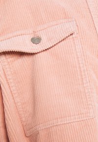 Jasnoróżowy materiał sztruksowy z pionowymi prążkami, wyposażony w kieszeń na piersi zabezpieczoną srebrnym zatrzaskiem oraz przeszyciami przy szwach.