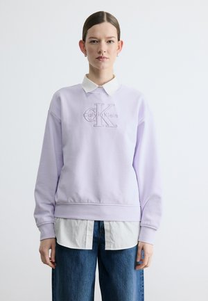 Femme portant un sweat-shirt Calvin Klein violet clair par-dessus une chemise blanche à col, avec un jean bleu, debout devant un fond blanc uni.