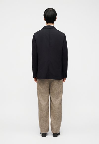 Άνδρας που στέκεται με την πλάτη γυρισμένη, φοράει μαύρο σακάκι, μπεζ παντελόνι φαρδιάς γραμμής και μαύρα παπούτσια σε απλό λευκό φόντο.