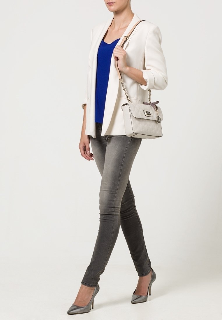 Femme portant un blazer blanc, un haut bleu, un jean skinny gris, des talons argentés et un sac à main beige avec une bandoulière en chaîne sur l’épaule.