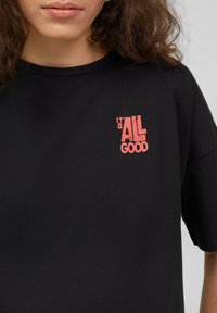 Zwarte katoen t-shirt met korte mouwen, voorzien van een rode grafische tekstafdruk met de tekst "het is ALL GOOD" op de bovenste linkerkant.