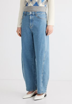 Osoba má na sebe svetlomodré široké džíny, biele topánky s hrotmi a sveter s dlhým rukávom s vzorom argyle.