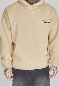 Hombre con sudadera beige con logo negro en el pecho y jeans gris oscuro, manos visibles con tatuajes, fondo neutro.