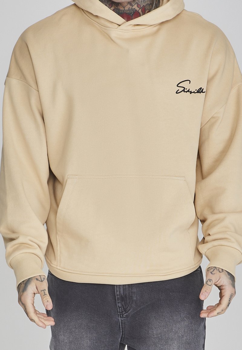 Hombre con sudadera beige con logo negro en el pecho y jeans gris oscuro, manos visibles con tatuajes, fondo neutro.