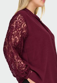 Top bordeaux con bottoni, realizzato in tessuto testurizzato, con maniche lunghe interamente in pizzo decorato con motivo floreale e paillettes per un tocco di dettaglio in più.