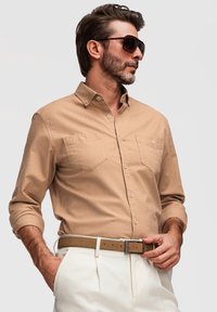 Homme avec barbe portant des lunettes de soleil, chemise beige à boutons avec manches retroussées, pantalon blanc et ceinture beige, posant avec une main dans la poche.
