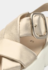 Δερμάτινο σανδάλι σε μεταλλικό χρυσό με χοντρή λευκή σόλα. Διαθέτει σταυρωτά λουριά και ορθογώνια αγκράφα. Ομαλή υφή.