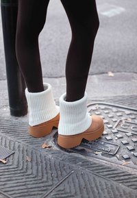 Botines tobilleros de punto acanalado en color crema con detalles en ante marrón y suelas plataforma, sobre un pavimento texturizado con hojas esparcidas.