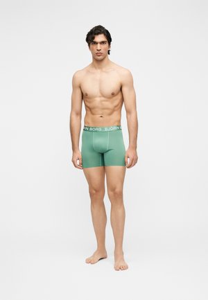 Mannelijk model staat blootsvoets tegen een witte achtergrond met groene Björn Borg boxershorts, kijkt recht vooruit met een neutrale uitdrukking.