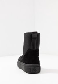 Botas negras hasta el tobillo con un superior sintético liso, tacón de ante texturizado y suela de goma. Incluye una lengüeta trasera para ponerlas fácilmente.