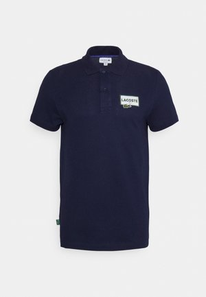 Laivastonsininen Lacoste-pikeepaita, jossa on lyhyet hihat, kaksinappinen nappilista ja suorakaiteen muotoinen logoplässi vasemman rinnan kohdalla.