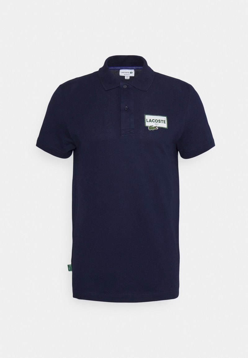 Polo Lacoste bleu marine à manches courtes, patte de boutonnage à deux boutons, et patch logo rectangulaire sur la poitrine gauche.