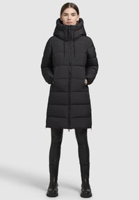 Abrigo de plumas largo negro con capucha, presenta un cierre de cremallera vertical, diseño acolchado y aberturas laterales. Combinado con leggins negros y botas.