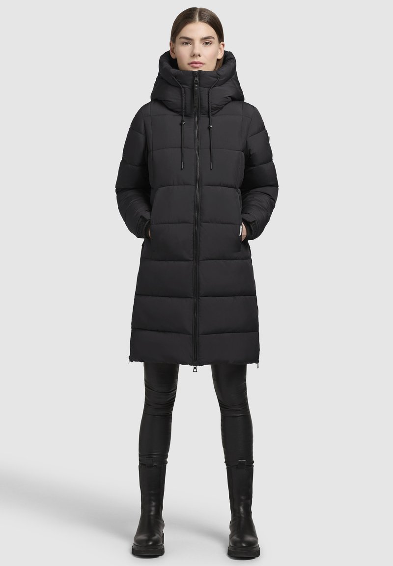 Abrigo de plumas largo negro con capucha, presenta un cierre de cremallera vertical, diseño acolchado y aberturas laterales. Combinado con leggins negros y botas.