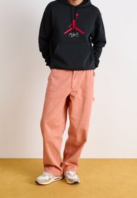 Sudadera negra con el logo rojo de Jordan, pantalones sueltos rosas y zapatillas beige con acentos rojos. Diseño simple, ajuste casual y tejido texturizado.