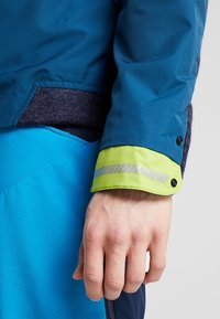 Main d'une personne près de la manche d'une veste bleue avec un poignet vert néon comportant une bande réfléchissante et des boutons-pression, sur fond gris clair.