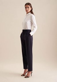 Biała koszula z długim rękawem i kołnierzykiem, zestawiona z granatowymi spodniami z wysokim stanem. Modelka stoi w czarnych sandałach na wysokim obcasie. Neutralne tło.