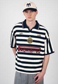 Jeune homme portant un polo rayé bleu marine et blanc avec le texte "PROHIBITED" et une casquette blanche avec un logo bleu foncé.