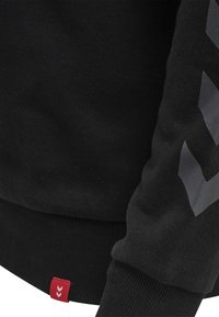 Schwarzer Sweatshirt mit strukturiertem Stoff, gerippten Bündchen und einem roten Etikett. Der Ärmel zeigt ein großes, graues Chevron-Muster.