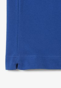 Blå pikétröja med en texturerad tyg, med rak fåll och sidslitsar. Stygnen är jämna längs kanterna.