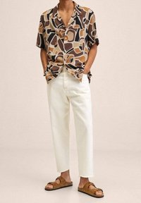 Person, der ein kurzärmliges Hemd mit abstraktem Muster, eine sandfarbene gerade Hose und braune Wildleder-Sandalen mit offenen Zehen trägt, steht mit den Händen in den Taschen.