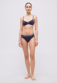 Γυναίκα που στέκεται ξυπόλυτη φορώντας μπλε σουτιέν και εσώρουχο με το λογότυπο "Polo Ralph Lauren" στη μέση, σε λευκό φόντο.