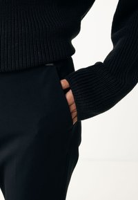 Suéter de punto negro acanalado con mangas largas, combinado con pantalones negros. La mano está colocada en el bolsillo lateral de los pantalones. Se observa una textura suave.