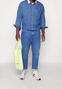 Jeansjacka med beige krage, ihop med denim jeans. Håller en ljusgrön totebag med "SAMS" tryckt. Neutralt färgade sneakers.