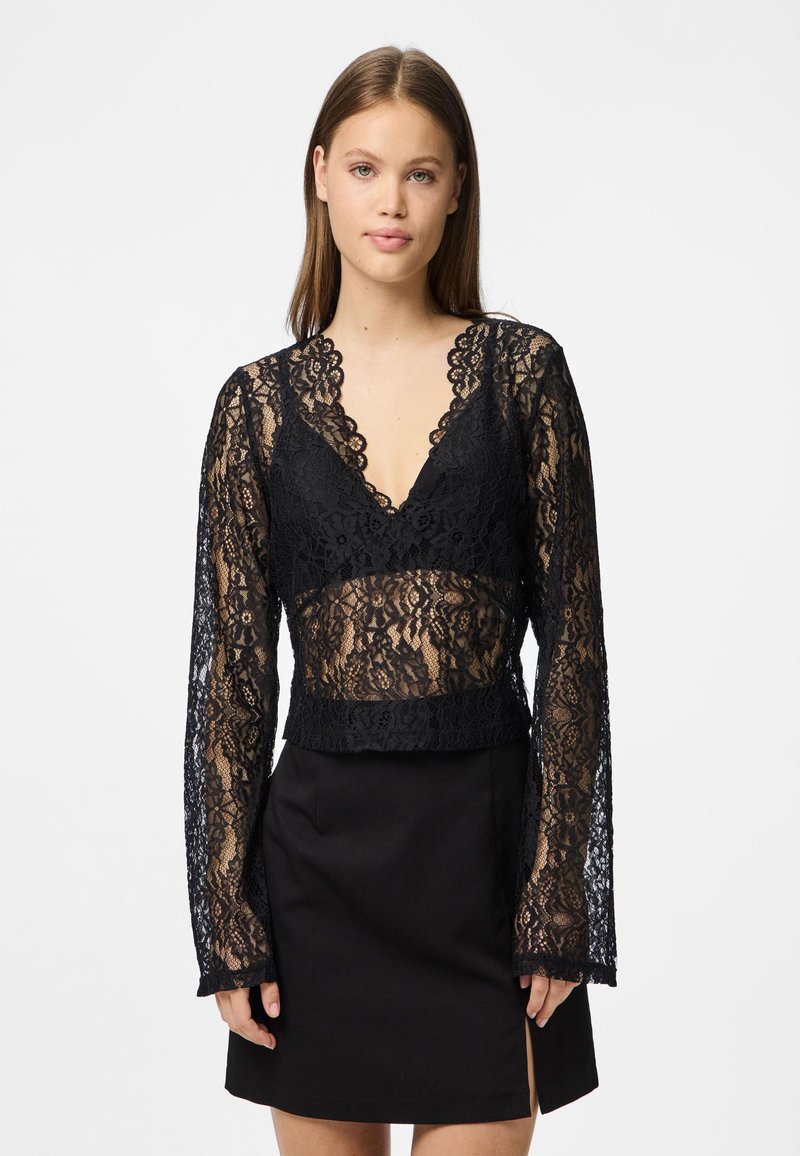 Crna čipkasta majica s dugim rukavima, dubokim V-izrezom, prozirnim dizajnom, oštrim rubovima i skraćenim fitom, kombinirana s crnom suknjom.