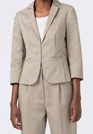 Personne à la peau foncée portant un blazer beige aux manches trois-quarts et un pantalon assorti sur un haut blanc.