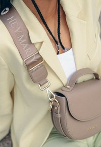 Sac en cuir beige en bandoulière avec une forme arrondie, quincaillerie dorée et une sangle amovible avec le texte "HEY MARY" en écriture légère.