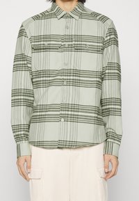 Camisa de cuadros verde hecha de tela suave con rayas negras horizontales y verticales, con dos bolsillos en el pecho y cierre de botones.