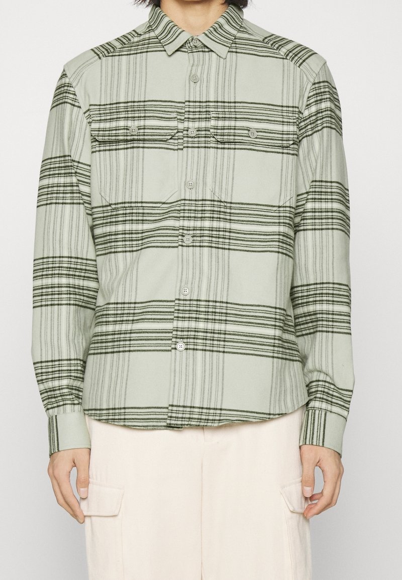Camisa de cuadros verde hecha de tela suave con rayas negras horizontales y verticales, con dos bolsillos en el pecho y cierre de botones.