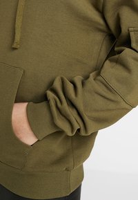 Sudadera con capucha color verde oliva en una mezcla suave de algodón, con bolsillo tipo canguro, puños acanalados y capucha con cordón. Tejido texturizado con un sutil grado de elasticidad.