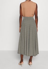 Femme portant un haut sans manches marron et une jupe midi plissée à imprimé géométrique taille haute, avec des sandales à talons beiges, se tenant de dos.