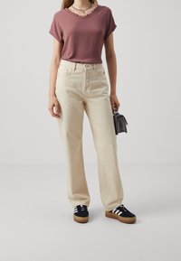 Beiga vida jeans i kombination med en mauve-blus med vågformad halsringning. Svarta sneakers med vita ränder och en liten svart handväska.