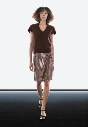 Brauner kurzärmeliger Strickpullover mit V-Ausschnitt, kombiniert mit einem roségoldenen Paillettenrock und schwarzen Sandalen mit Absatz, vor neutralem Hintergrund präsentiert.