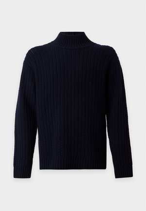 Homme portant un pull en maille bleu marine foncé, un pantalon gris à jambes larges et des chaussures noires à lacets, debout devant un fond blanc uni.