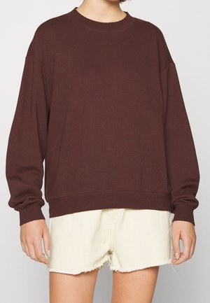Person iført en løs, langærmet mørkebrun sweatshirt kombineret med lyse cremefarvede shorts med frynset kant mod en ensfarvet baggrund.