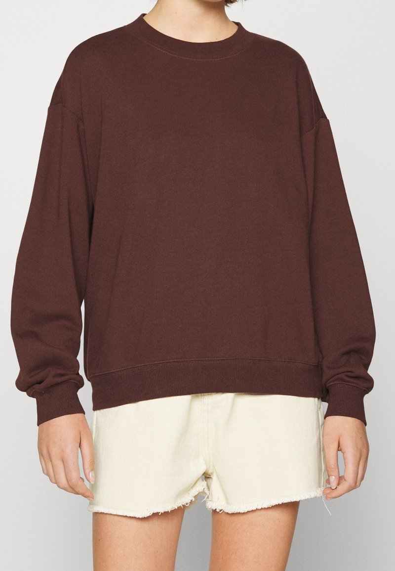 Personne portant un sweat-shirt ample à manches longues marron foncé associé à un short crème clair à ourlet effiloché sur un fond uni.
