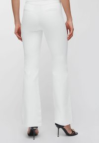 Donna che indossa pantaloni a zampa bianchi aderenti e sandali neri con tacco alto e decorazioni, vista da dietro, su uno sfondo semplice.