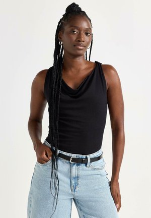 Kvinde med langt flettet hår iført en sort ærmeløs top og lyseblå jeans, stående med den ene hånd i lommen mod en hvid baggrund.