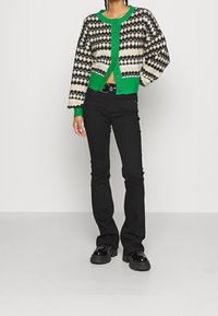 Černý pletený kardigan s bílými a zelenými diamantovými vzory, zelenými žebrovanými okraji a knoflíky, doplněný černými přiléhavými kalhotami a robustními botami.