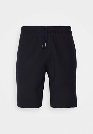 Mies, jolla on sininen kukallinen nappipaita, mustat shortsit, valkoiset sukat ja valkoiset lenkkarit, seisoo kädet taskuissa.