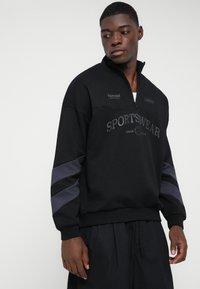 Felpa nera con colletto a zip a quarto, con la scritta "SPORTSWEAR" in grigio scuro e strisce orizzontali grigie sulle maniche.