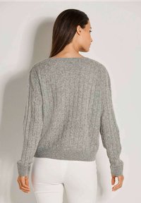 Frau mit langen dunklen Haaren, die einen grauen Strickpullover und weiße Hosen trägt, steht mit dem Rücken zur Kamera vor einer einfachen Wand.